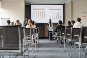 Two women leading a corporate training. They stand holding microphones in front of a projector screen addressing an audience.