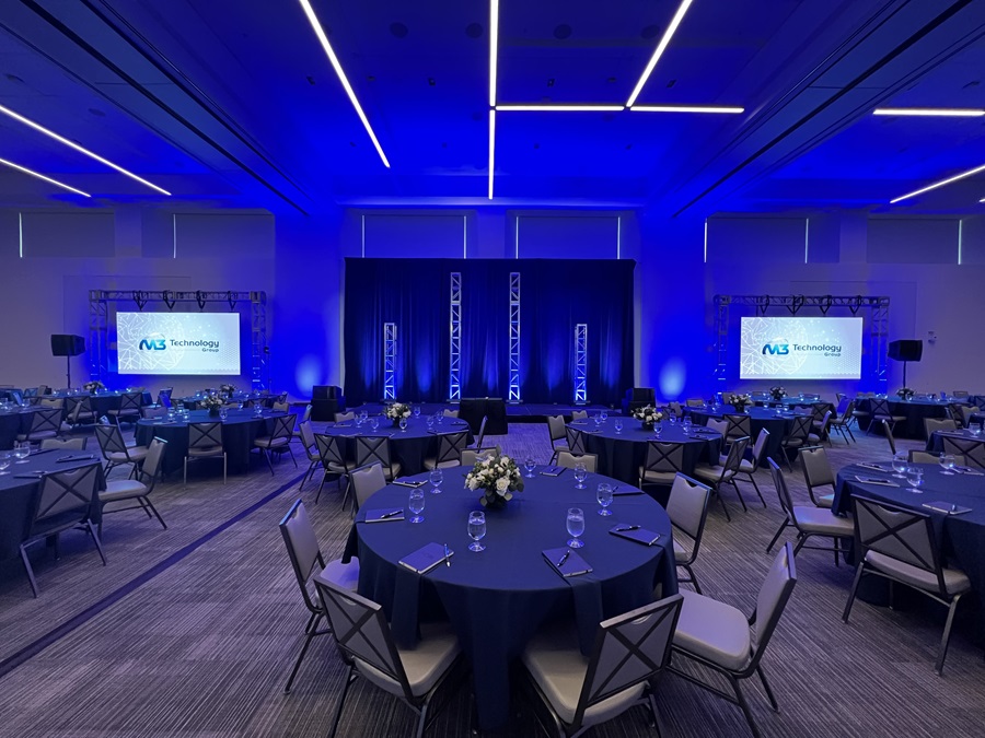 A professionally staged event space set up by M3 Technology Group, featuring round tables with blue tablecloths, floral centerpieces, and two large projection screens displaying the M3 Technology Group logo under ambient blue lighting.