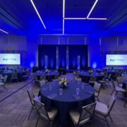 A professionally staged event space set up by M3 Technology Group, featuring round tables with blue tablecloths, floral centerpieces, and two large projection screens displaying the M3 Technology Group logo under ambient blue lighting.