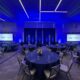 A professionally staged event space set up by M3 Technology Group, featuring round tables with blue tablecloths, floral centerpieces, and two large projection screens displaying the M3 Technology Group logo under ambient blue lighting.