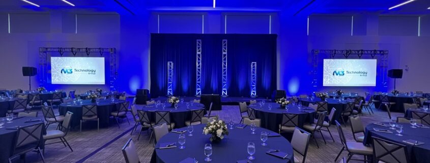 A professionally staged event space set up by M3 Technology Group, featuring round tables with blue tablecloths, floral centerpieces, and two large projection screens displaying the M3 Technology Group logo under ambient blue lighting.