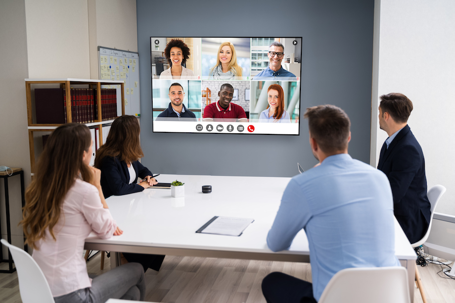 A conference room with employees gathered around a video display that shows six remote team members. 