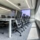 A conference room setup featuring a long table, chairs, and a large screen display at the end and a wall of windows to the right with motorized shades lowered halfway down.