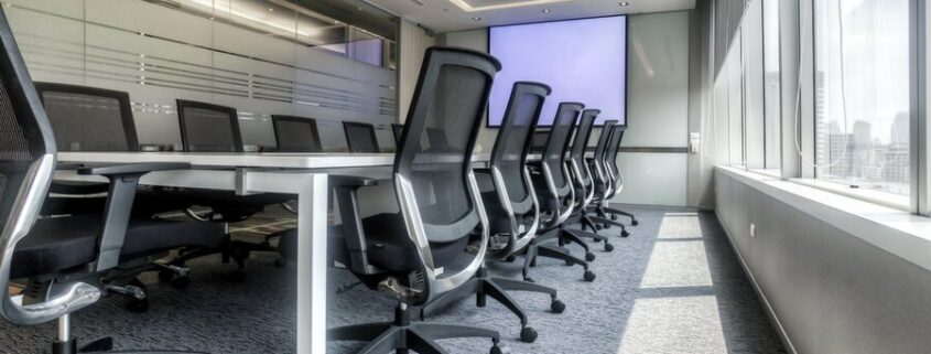 A conference room setup featuring a long table, chairs, and a large screen display at the end and a wall of windows to the right with motorized shades lowered halfway down.