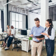 Two people looking at an open folder in an office setting with tables, chairs, and computers.