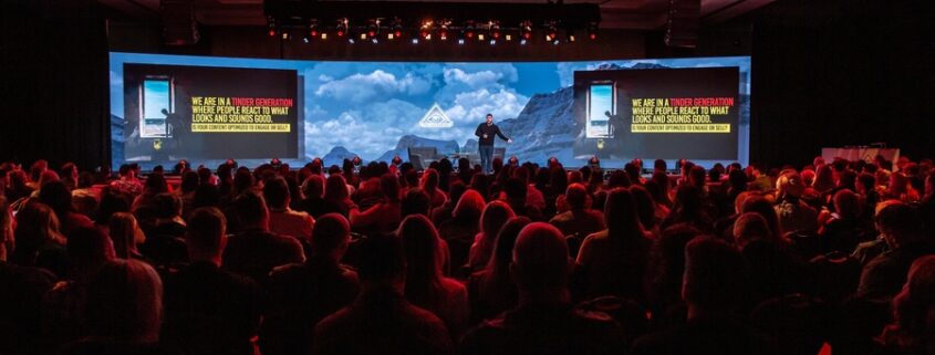 A person on a stage in front of a live audience with a display behind them and red lighting illuminating the crowd.