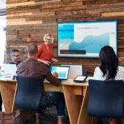 A huddle room with one person standing at a display and five sitting at a table with laptops, tablets, and controllers.