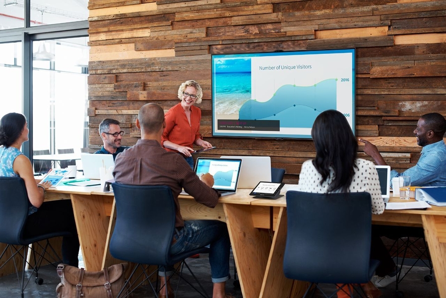 A huddle room with one person standing at a display and five sitting at a table with laptops, tablets, and controllers.