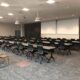 A commercial training room featuring rows of rectangular tables with two large projector screens at the front of the room.