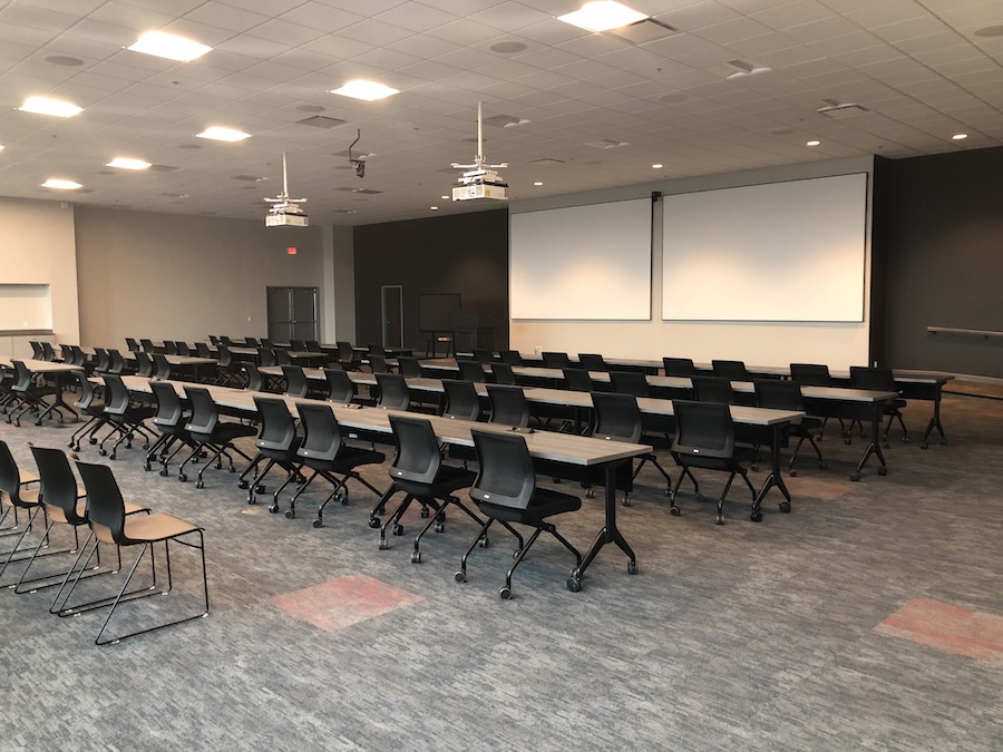 A commercial training room featuring rows of rectangular tables with two large projector screens at the front of the room.